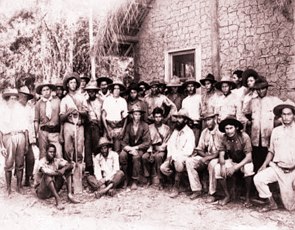 Foto antigo de vários homens na frente de uma casa de barro. Os homens usam chapéus.