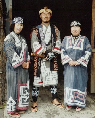 Foto de um homem e duas mulheres ainus com vestimenta tradicional do seu povo.