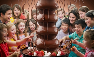 Imagem de crianças e adultos comendo chocolate numa torre de chocolate derretido.