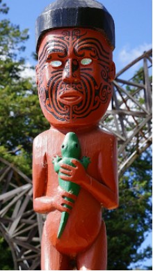 Foto de uma escultura maori representando um homem maori com tatuagem no rosto e segurando um jacar&eacute; pequeno.