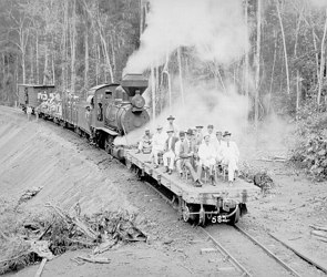 Foto em preto e branco de uma locomotiva soltando fumaça e pessoas na frente dela