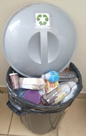 Foto de uma lixeira com vários materiais recicláveis dentro como garrafa de vidro, caixa de ovos, lata de refrigerante e embalagem de vinagre