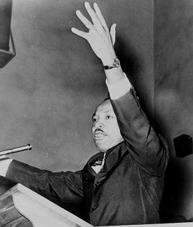 Foto em preto e branco de um homem negro de bigode fazendo um comício com o braço esquerdo levantado.