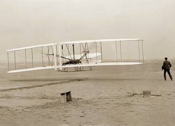 Foto em preto e branco mostrando um pequeno avi&atilde;o levantando voo e um homem ao lado