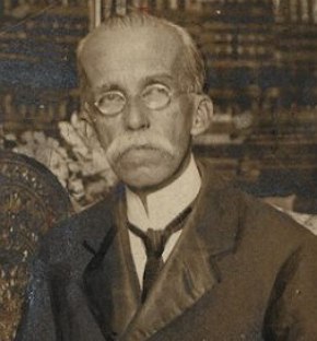 Foto em preto e branco amarelada mostrando homem idoso, calvo, usando óculos e de bigode branco